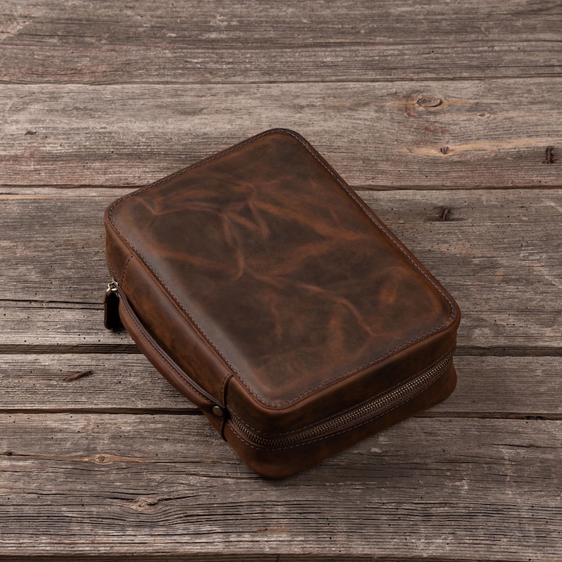 May include: A rectangular brown leather travel case with a smooth, distressed finish and a wrap-around zipper closure. The case features a sturdy side handle and rounded corners, showcasing a deep, rich brown color with natural variations.