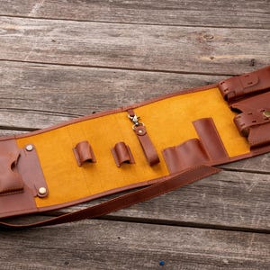 K&ouml;nnte beinhalten: Eine braune Werkzeugrolle aus Leder mit leuchtend gelbem Innenfutter. Der Organizer verf&uuml;gt &uuml;ber mehrere Taschen, zwei kleine Schlaufen, einen drehbaren Metallverschluss an einem Lederriemen und zwei gr&ouml;&szlig;ere Taschen mit Druckknopfverschl&uuml;ssen.