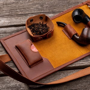 K&ouml;nnte beinhalten: Eine braune Tabakpfeifentasche aus Leder mit senfgelbem Innenfutter, einer kleinen Lederschale mit losem Tabak, einer Ledertasche f&uuml;r Zubeh&ouml;r sowie zwei Holzpfeifen, die mit Lederriemen und Metallbeschl&auml;gen sicher befestigt sind.