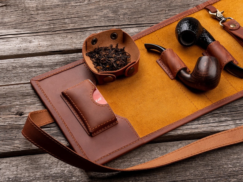 K&ouml;nnte beinhalten: Eine braune Tabakpfeifentasche aus Leder mit senfgelbem Innenfutter, einer kleinen Lederschale mit losem Tabak, einer Ledertasche f&uuml;r Zubeh&ouml;r sowie zwei Holzpfeifen, die mit Lederriemen und Metallbeschl&auml;gen sicher befestigt sind.