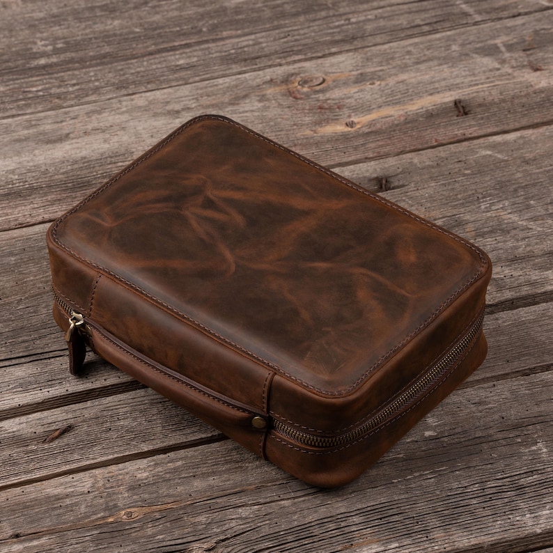 May include: A rectangular brown leather case with a smooth, distressed finish and visible grain texture. The case features a wrap-around metal zipper closure with a matching leather pull tab and a side handle attached with a small metal rivet.