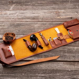 K&ouml;nnte beinhalten: Eine braune Tabakpfeifentasche aus Leder mit gelbem Innenfutter, die zwei Holzpfeifen, ein Metallwerkzeug, eine kleine Lederschale mit losem Tabak, eine Metalldose und eine wei&szlig;e Reinigungsb&uuml;rste enth&auml;lt, gesichert mit einem langen Lederriemen.