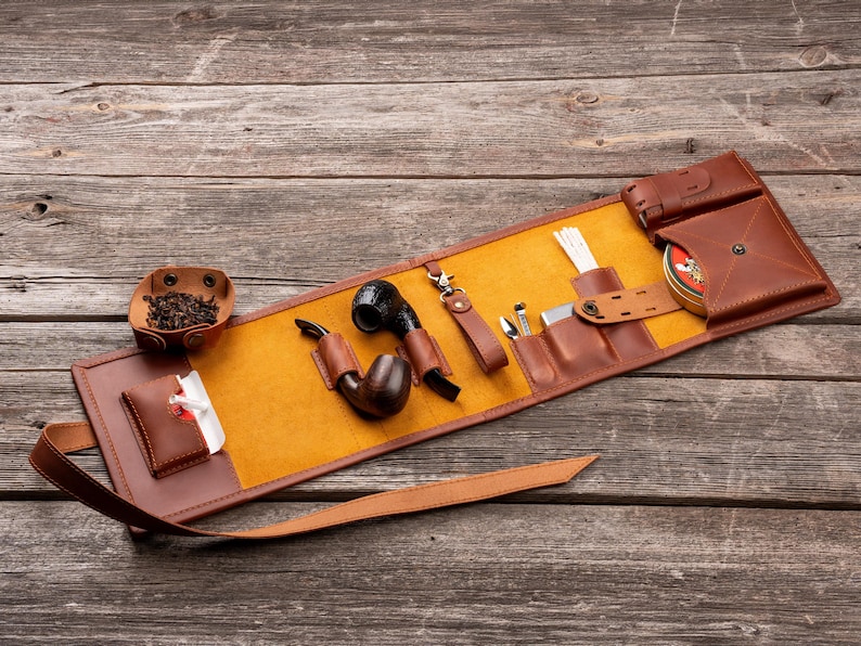 K&ouml;nnte beinhalten: Eine braune Tabakpfeifentasche aus Leder mit gelbem Innenfutter, die zwei Holzpfeifen, ein Metallwerkzeug, eine kleine Lederschale mit losem Tabak, eine Metalldose und eine wei&szlig;e Reinigungsb&uuml;rste enth&auml;lt, gesichert mit einem langen Lederriemen.