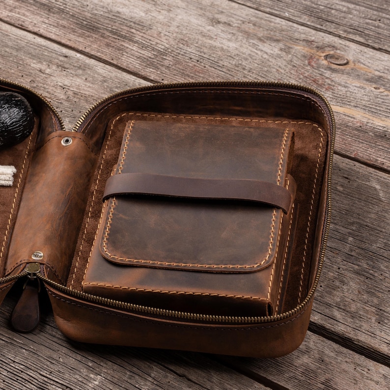 May include: A brown leather zippered case featuring a rectangular interior compartment secured by a horizontal leather strap. The case has visible stitching along the edges and a smooth, matte finish, designed to hold items in a structured, organized manner.