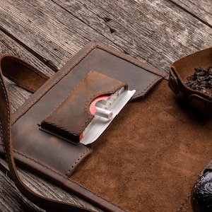 K&ouml;nnte beinhalten: Ein brauner Tabakbeutel aus Leder mit einer speziellen Tasche f&uuml;r eine Zigarettenschachtel, einer kleinen Druckknopfschale mit losem Tabak und einer Lederschlaufe zur Aufbewahrung einer Pfeife, alles mit sichtbaren N&auml;hten versehen.