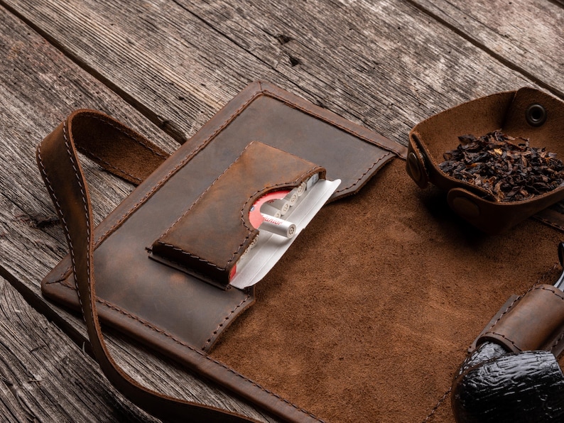 K&ouml;nnte beinhalten: Ein brauner Tabakbeutel aus Leder mit einer speziellen Tasche f&uuml;r eine Zigarettenschachtel, einer kleinen Druckknopfschale mit losem Tabak und einer Lederschlaufe zur Aufbewahrung einer Pfeife, alles mit sichtbaren N&auml;hten versehen.