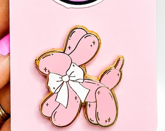 May include: A gold-toned metal enamel pin shaped like a pink balloon dog with a white bow, attached to a light pink display card. Above the pin is a circular sticker featuring a black and pink graphic with the text MIND THE NEEDLE.