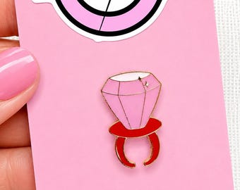 May include: A pink enamel pin shaped like a ring pop candy with a faceted light pink diamond top and a red band. The pin is attached to a pink display card featuring a circular logo with the text MIND THE NEEDLE and a needle graphic.