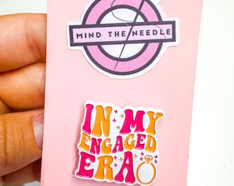 May include: Pink card with two stickers. The top sticker has a pink circle with a needle and thread design and the text "MIND THE NEEDLE". The bottom sticker says "IN MY ENGAGED ERA" with a ring graphic.