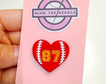 May include: A pink card with two stickers. The top sticker is a pink circle with a needle and thread design and the text "MIND THE NEEDLE". The bottom sticker is a red heart with a baseball design and the number "87".