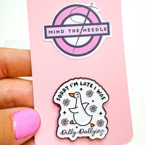 May include: A pink card with two enamel pins. The top pin reads "Mind the Needle" in a black and white design. The bottom pin features a white goose with the text "Sorry I'm Late I Was Dilly Dallying" and daisy accents.