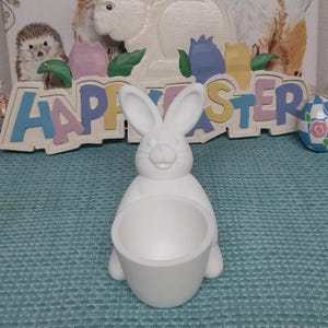 May include: White ceramic bunny rabbit planter with a round pot. The bunny is sitting on a blue and white patterned surface. The bunny has a happy expression and is holding a pot in its front paws.