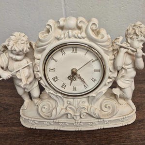 Op de afbeelding: Crèmekleurige decoratieve klok met een ronde wijzerplaat en Romeinse cijfers. Twee cherubijnen flankeren de klok, een die viool speelt en de ander fluit. De klok heeft een gedetailleerd ontwerp en een quartz uurwerk.