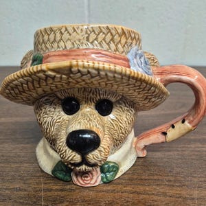 Puede incluir: Una taza de cerámica con forma de oso de peluche que lleva un sombrero de paja con una cinta rosa y una flor azul. El oso tiene ojos negros y una nariz negra. El asa tiene forma curva con un color rosa y marrón.