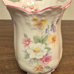 Puede incluir: Un jarrón de cerámica blanco con borde festoneado, adornado con un estampado floral con flores blancas, rosas, amarillas y azules. El borde está delineado con un ribete rosa. El jarrón tiene una forma ligeramente acampanada y es para exhibir flores.