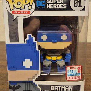 May include: A boxed Funko Pop! 8-bit Batman vinyl figure. The figure features a pixelated design in blue, grey, and yellow. The box includes the DC Super Heroes logo and a "Fall Convention Exclusive" sticker.