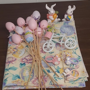 May include: A collection of Easter decorations including a white bicycle, pink and white bunny napkin rings, pink and white ceramic bunnies, and pink and white Easter eggs on sticks.