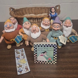 Op de afbeelding: Een verzameling Sneeuwwitje en de zeven dwergen knuffelfiguren, een klein kartonboekje getiteld Snow White and the Seven Dwarfs, een stickervel en een gevlochten rieten mand. De figuren hebben verschillende gekleurde mutsen en tunieken.