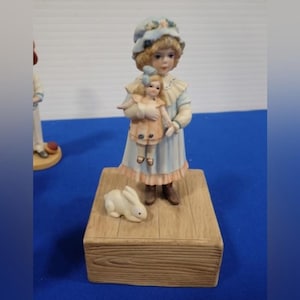 Könnte beinhalten: Eine Porzellanfigur eines jungen Mädchens in einem blauen Kleid, das eine Puppe hält. Sie steht auf einem Holzsockel mit einer weißen Hasenfigur.