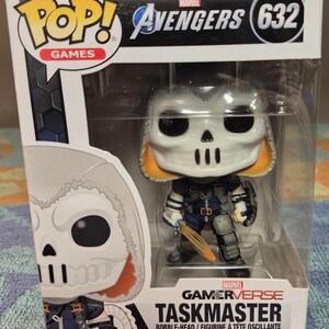 May include: A Taskmaster bobblehead figure from the Marvel Gamerverse series. The figure has a white skull-like mask, orange accents, and is holding a sword. The packaging includes the Avengers logo and the number 632.