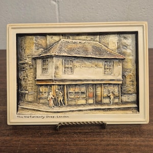 May include: A rectangular wall plaque featuring a relief sculpture of The Old Curiosity Shop in London. The cream-coloured frame surrounds a detailed scene of the building with figures, windows, and the text Old Curiosity Shop and The Old Curiosity Shop. London.