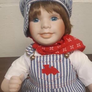 Peut inclure: Une poupée en porcelaine vêtue d'une salopette rayée bleu et blanc avec un bandana rouge et une casquette rayée bleu et blanc. La poupée a les yeux bleus et un visage souriant.