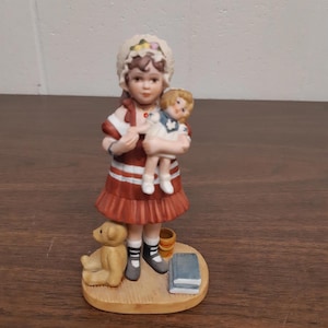 Könnte beinhalten: Eine Keramikfigur eines jungen Mädchens in einem roten Kleid und einer weißen Haube, das eine Puppe hält. Ein Teddybär, ein kleiner Fass und ein Buch befinden sich zu ihren Füßen. Die Figur steht auf einem Holzsockel.