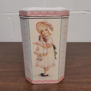 Könnte beinhalten: Vintage Metall-Aufbewahrungsdose mit rosa-weißem Deckel und dekorativer Illustration. Die Illustration zeigt ein junges Mädchen mit Hut, das eine Puppe hält. Die Dose hat ein hellrosa und blaues Rautenmuster an den Seiten.