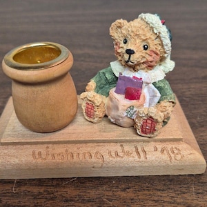 May include: A figurine of a light brown teddy bear wearing a white bonnet and green coat, sitting next to a small wooden pot with a gold rim. The items are mounted on a rectangular wooden base with the text Wishing Well '98 carved into the front.