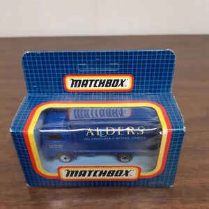 Pode incluir: Um caminhão de brinquedo azul Matchbox com as palavras "Alders All Together A Better Choice" no lado. O caminhão está em uma vitrine de plástico transparente com um fundo de papelão azul e amarelo.
