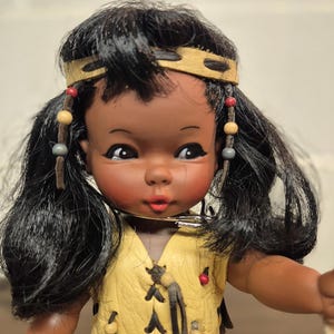 May include: A vintage doll with dark skin, black hair, and a yellow leather-like vest. The doll has a headband and beaded accents in her hair. The vest is adorned with decorative stitching and small red and blue accents. The doll's face has rosy cheeks and a small red mouth.