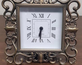 May include: An ornate gold-toned clock with a white face and black Roman numeral hour markers. The clock is in a decorative frame with a scrolled design.