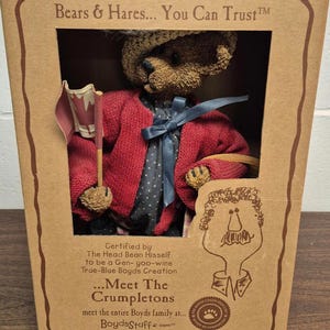 May include: A teddy bear doll in a red cardigan, blue bow, and hat, holding a small flag, presented in a cardboard box. The box features text including "Bears & Hares... You Can Trust" and "Meet The Crumpletons".