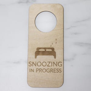 May include: A wooden door hanger with a cutout for a door knob. The hanger is light brown and has the text "Snoozing in Progress" and a stylized image of a bed with Z's above it.