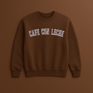 May include: A brown crewneck sweatshirt with the text "CAFE CON LECHE" in white, arched across the chest. The sweatshirt is a solid, warm brown color, with a ribbed neckline, cuffs, and hem. The background is a matching brown.