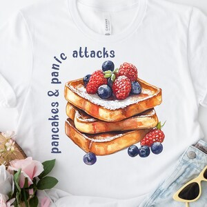 Könnte beinhalten: Weißes T-Shirt mit einem Grafikdruck von einem Stapel French Toast, der mit Blaubeeren und Himbeeren belegt ist. Der Text "pancakes & pan/c attacks" ist auf das Shirt gedruckt.