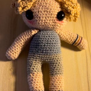 May include: A crocheted doll with blonde hair, blue and pink clothing, and black shoes. The doll has large black eyes and a pink blush.