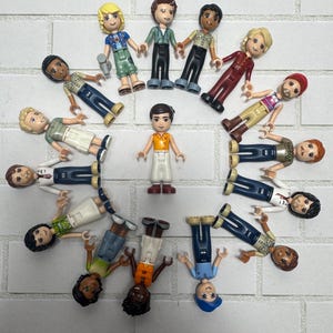 May include: A collection of Lego mini-figures arranged in a circle on a white brick surface. The figures have various skin tones, hairstyles, and outfits, including shirts, pants, and accessories. One figure stands in the center.
