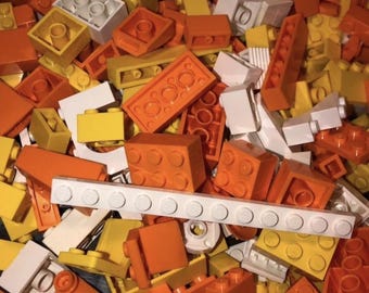 May include: A pile of colorful plastic building blocks in shades of orange, yellow, and white. The blocks are various shapes and sizes, including rectangular bricks and square blocks with circular studs. A long white block with circular studs is also visible.
