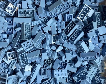 May include: A large collection of light gray plastic building bricks in various shapes and sizes. The pile includes rectangular blocks with multiple studs, flat plates, and specialized pieces, all featuring a uniform matte finish and hollow undersides.