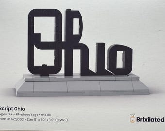 May include: A black brick-built model spelling the word Ohio in a script font, mounted on a light gray rectangular base. The text on the image includes Script Ohio, Ages 7+, 89-piece Lego model, Item # MCB003, Size 5" x 1.9" x 3.2" (LxWxH), and Brixilated.