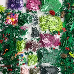 May include: Assortment of small plastic bags filled with various colors of Lego pieces, including green, red, purple, pink, and black. The pieces are in the shape of leaves, flowers, and other plant parts. The bags are arranged in a grid pattern on a white surface.