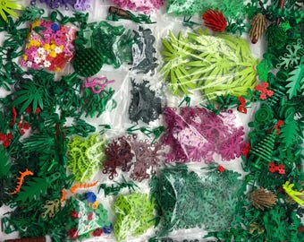 May include: Assortment of small plastic bags filled with various colours of Lego pieces, including green, red, purple, pink, and black. The pieces are in the shape of leaves, flowers, and other plant parts. The bags are arranged in a grid pattern on a white surface.