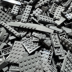Puede incluir: Una gran colección de ladrillos de construcción de plástico gris de diversas formas y tamaños. El montón incluye placas rectangulares, piezas cuadradas y vigas largas, todas con los característicos pernos circulares en la parte superior.