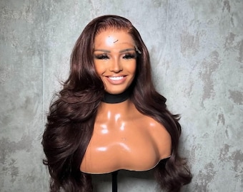 May include: A dark brown wig with long, wavy layers is displayed on a mannequin head. The wig features a natural-looking hairline and is styled with voluminous curls. The mannequin is mounted on a black stand against a gray textured background.