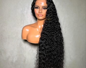 May include: A mannequin head with a long, curly black wig. The wig has a natural-looking hairline and is styled with voluminous, cascading curls. The mannequin has a tan complexion, with painted facial features, including eyelashes and eyebrows. The background is a plain gray wall.