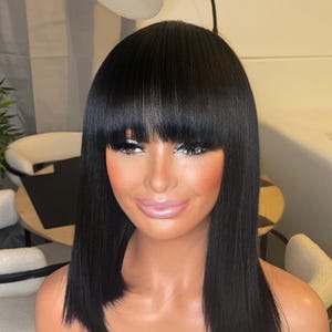 Peut inclure: Une perruque noire avec une coupe au carré et une frange. La perruque est coiffée avec des cheveux raides et lisses. La perruque est sur une tête de mannequin avec un teint clair et du maquillage.