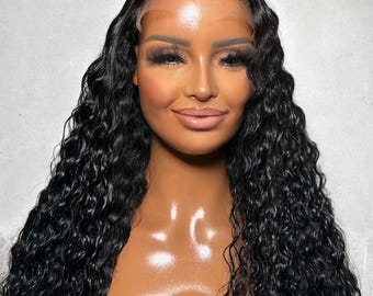 Può includere: Una donna con lunghi capelli neri ondulati. I capelli sono acconciati con un'attaccatura naturale e una scriminatura laterale profonda. La donna ha un trucco completo, comprese ciglia scure e un rossetto nude. Lo sfondo è grigio chiaro.