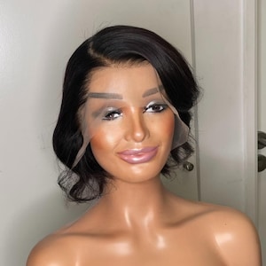 Peut inclure: Une perruque noire courte avec une frange en dentelle, coiffée en vagues lâches. La perruque est sur une tête de mannequin avec un teint clair.