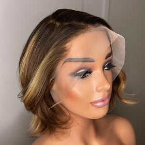 Peut inclure: Une perruque courte et ondulée, de couleur brune avec des mèches blondes. La perruque est coiffée avec une raie sur le côté et a une frange en dentelle.